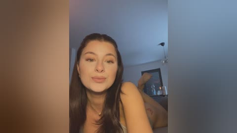Video of a young woman with long dark hair, fair skin, and subtle makeup, posing seductively in a modern, dimly lit bedroom with minimalistic decor.