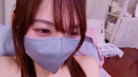 Video of a young East Asian woman with long brown hair, wearing a light blue face mask, sitting in a cozy, cluttered room with white shelves and a pink blanket.