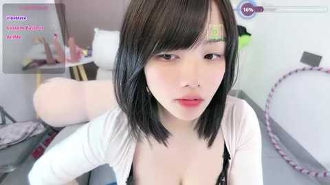 A video of a young East Asian woman with shoulder-length black hair, wearing a white cardigan, sitting on a bed with a pink hoop. The room is sparsely decorated with a pink teddy bear and a green lamp.