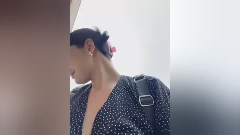 Video of a woman with dark hair tied back, wearing a black polka-dot dress with a deep V-neck and a black leather shoulder bag. The background is blurred, creating a soft focus effect.