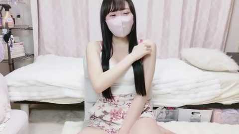 Video of a young Asian woman with long black hair, wearing a floral dress, face mask, and gloves, seated on a bed with white linens, in a soft-lit room with pastel curtains.