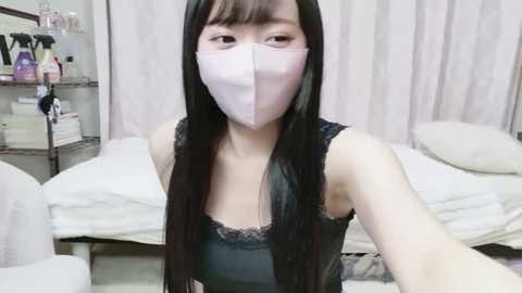 A video of an East Asian woman with long black hair, wearing a light pink face mask, a black lace camisole, and a white towel, posing in a bedroom with white curtains and a bed.