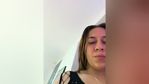 Video of a young Latina woman with light brown skin, straight brown hair, wearing a black top, blowing air at a camera lens, taken indoors with white walls and minimal decor.