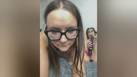 A video of a woman with glasses, light skin, and wet hair, in a gray top, holding a black dildo, in a dimly lit room.