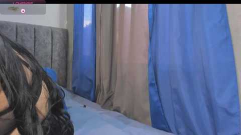 A video of a bedroom with beige and blue curtains, a gray upholstered headboard, and a bed covered in white sheets. The room appears to be in a state of disarray.