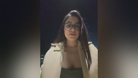 Video of a dark-haired woman with glasses and a beige coat, standing against a dark, blurry background.
