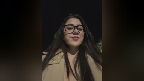 Video of a young woman with long dark hair, wearing black-framed glasses and a beige coat, smiling softly against a dark background.