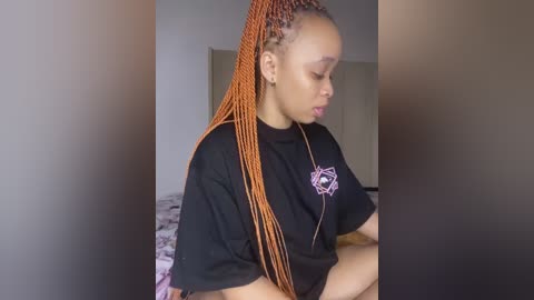 Video of a young Black woman with braided hair, wearing a black T-shirt with a white graphic, seated indoors.
