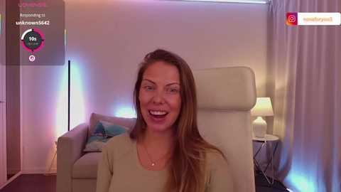 Video of a smiling, fair-skinned woman with long brown hair, wearing a beige top, sitting in a beige armchair in a softly lit, modern living room with a white lamp on a side table and a blue cushion on a sofa.