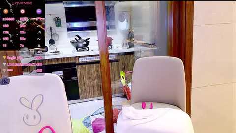 Video of a modern kitchen with a white chair, light grey cushion, and pink towel, featuring a wall-mounted TV displaying cooking recipes, wooden cabinets, and stainless steel appliances.
