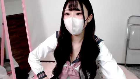 Video of an Asian woman with long black hair, wearing a Japanese schoolgirl outfit, white mask, and standing in a room with a pink-framed mirror and a metal folding chair.
