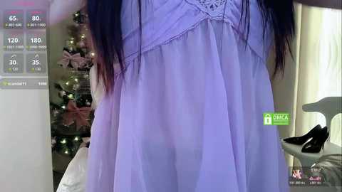 A video showing a woman in a sheer, light purple nightgown, with long dark hair, standing in a bedroom with a Christmas tree and a white chair in the background.