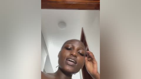 Video of a dark-skinned Black woman with short hair, slightly smiling, adjusting her hair with her right hand. Background shows a white ceiling and wooden trim.