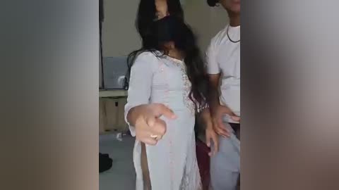 Media: Video of a young woman in a white, floral-patterned dress with long sleeves and a high neckline, her long, dark hair partially covering her face, gesturing with her right hand. A man in a white t-shirt stands beside her. Background includes a blurred TV and beige walls.