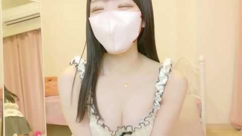 Video of an Asian woman with long black hair, wearing a white face mask, frilly top, and a small necklace, indoors, smiling, in a softly lit room.