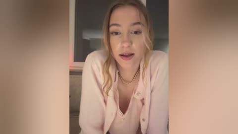 Video of a blonde Caucasian woman with fair skin, wearing a light pink blazer and necklace, looking directly at the camera with a surprised expression, in a dimly lit room.