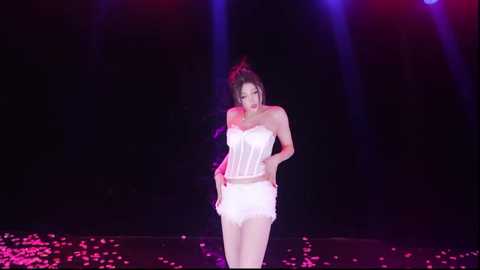 Video of an Asian woman with dark hair in a bun, wearing a white corset and fringed skirt, standing on a stage littered with pink rose petals under purple and red stage lights.