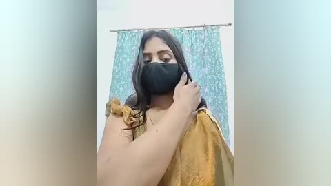 Video of a woman with medium skin tone, long dark hair, wearing a black face mask and a mustard-yellow sari, adjusting her hair in front of a blue floral-patterned shower curtain.