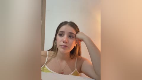 Video of a young Caucasian woman with straight, light brown hair, wearing a yellow and white spaghetti-strap top, leaning on a white wall, looking contemplative.