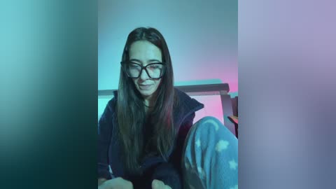 Video of a young woman with long brown hair, wearing glasses, a dark jacket, and light blue pajama pants, seated in a room with a purple and blue color gradient background.