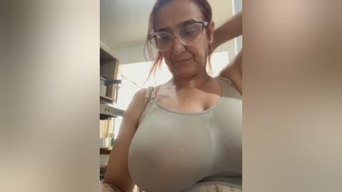 Video of a light-skinned woman with red hair in glasses, wearing a tight, light-colored top, standing in a dimly lit room with shelves and books in the background.