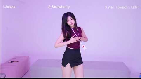 A video of an East Asian woman with long black hair, wearing a sleeveless maroon crop top and black mini-skirt, standing in a minimalist, white-walled room.