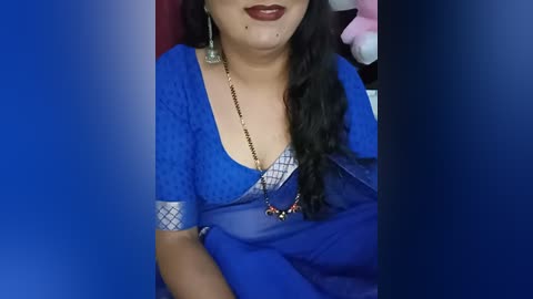 Video of a woman with light brown skin and long, wavy black hair, wearing a blue saree with a diamond-shaped pattern and a gold necklace, seated on a chair with a blurred background.
