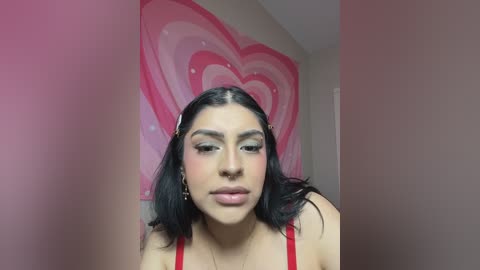 A video of a young woman with medium skin tone, long black hair, wearing a red tank top, standing in front of a pink heart wall, with a soft-focus background.