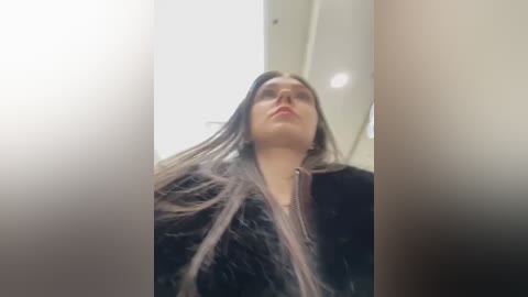 A video of a young woman with long, straight dark hair, wearing glasses and a black jacket, taken from a low angle, emphasizing her facial features and hair.