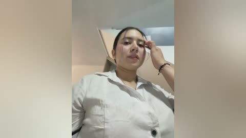 Video of a young Asian woman with light skin, dark hair, and wearing a white button-up shirt. She has blush marks on her cheeks, and one hand is touching her face. The background is a plain, beige wall.