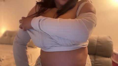 Video of a woman with medium brown skin, long dark hair, wearing a gray off-shoulder crop top and striped pajama bottoms, hugging herself in a softly lit, beige-toned living room with a couch in the background.