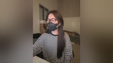 Video of a young Asian woman with long black hair, wearing black-framed glasses, a black mask, and a black-and-white striped shirt, sitting in a dimly lit living room with beige walls, a window, and a laundry basket in the background.
