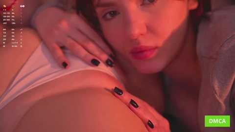 Video of a young woman with short, curly hair, wearing a white shirt, lying on a bed. Her hand with black nails touches her face. Red light casts a warm glow over the scene.
