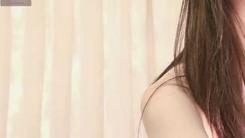 Video of a woman's shoulder and neck, showing long, wet brown hair cascading down. She is wearing a white, see-through top. Background features vertical, cream-colored, semi-transparent curtains.