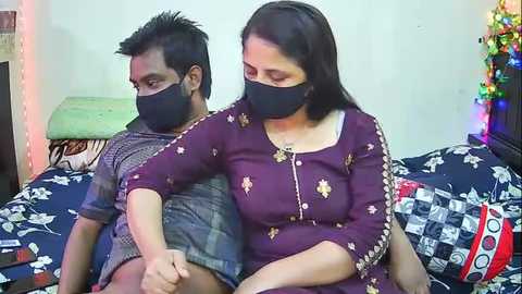 Video of an Indian couple wearing black masks, sitting on a bed with patterned blankets. The woman has shoulder-length dark hair, medium build, and a maroon kurta with gold embroidery. The man has a short beard and medium build, wearing a grey t-shirt.