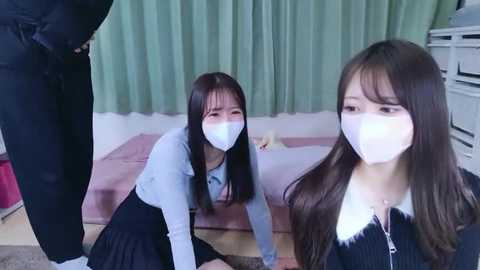 Video of three Asian women, two in light blue masks and black skirts, kneeling on a pink bed in a bedroom with green curtains and white walls.
