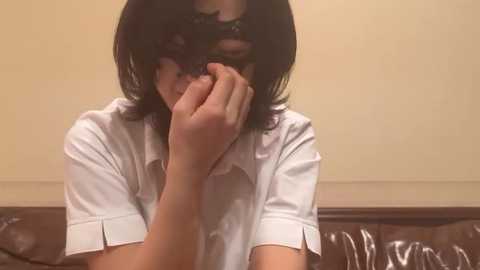 Video of a person with shoulder-length black hair, wearing a white button-up shirt, hiding their face with their hand, sitting on a brown leather couch.