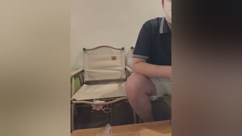 Video of a person in a dark polo shirt and white shorts, seated on a beige folding chair in a minimalist, beige-walled room with a wooden table visible in the foreground.
