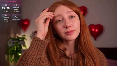 Video of a young woman with long red hair, wearing a brown knitted sweater, indoors with red heart decorations.