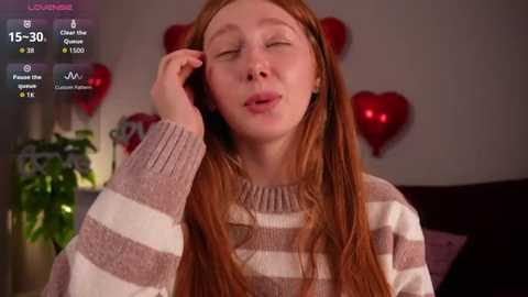 Video of a young woman with long red hair, wearing a striped sweater, closing her eyes and touching her face. Background shows a heart-shaped light and a potted plant.