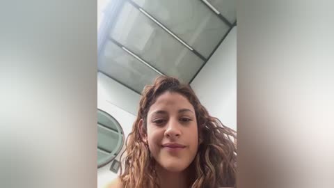 Video of a young Latina woman with curly brown hair, light skin, and a gentle smile, standing in a modern room with a glass ceiling and metallic accents.
