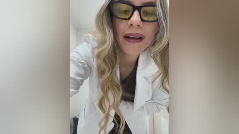 Video of a young, fair-skinned woman with long, wavy blonde hair, wearing large, black-rimmed glasses and a white lab coat, standing in a dimly lit room with blurred beige walls.