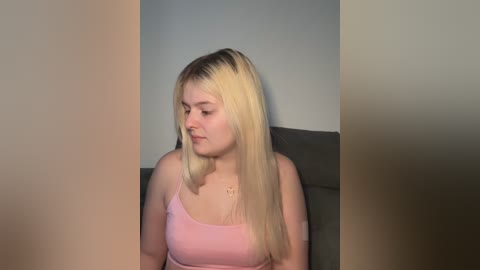 A video of a young, fair-skinned woman with long, straight, platinum blonde hair, wearing a pink spaghetti strap tank top, seated against a plain gray wall, looking down.