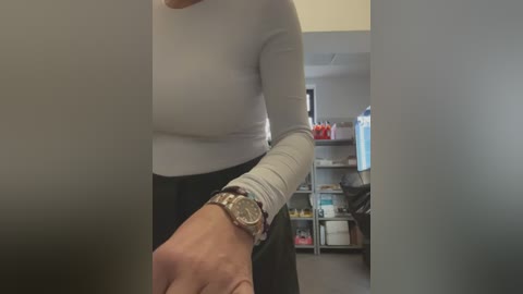Video of a woman with light skin, wearing a white long-sleeve top and black pants, standing in an office. She has a silver wristwatch on her left wrist, with a blurred background featuring shelves and a desk.