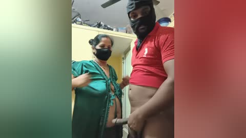 Video of a woman in a teal dress and mask, standing beside a man in a red shirt and mask holding a dildo. Both are indoors in a cluttered room.