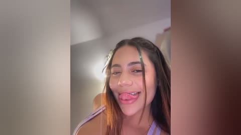 Video of a young woman with medium skin tone, straight brown hair, and green hair clips, sticking out her tongue while wearing a light-colored top, against a blurred indoor background.