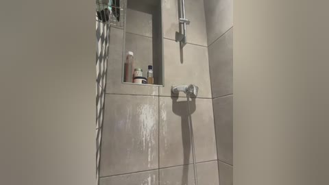 A video of a modern bathroom shower with beige tiles, a chrome showerhead, and a built-in shampoo and soap dispenser. Shampoo bottles are visible in the recessed shelf. The image has a neutral, clean aesthetic.