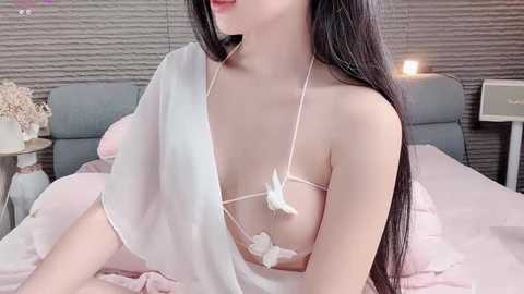 Video of a fair-skinned woman with long black hair, wearing a white lace bralette with pink flowers, draped in a sheer white fabric, sitting on a bed with a grey headboard in a minimalist room.