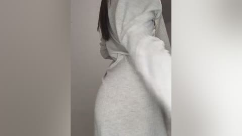 A video of a woman in a light grey sweatshirt and leggings, standing with her back to the camera, showing her side profile and long dark hair. The background is a plain, off-white wall.