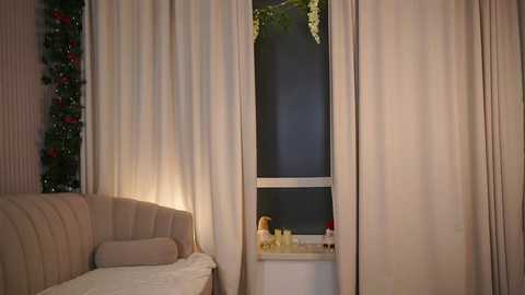 Video of a cozy, dimly-lit living room with beige curtains covering a window. A cream sofa with a decorative pillow sits to the left. Christmas garland with red ornaments hangs on the wall.